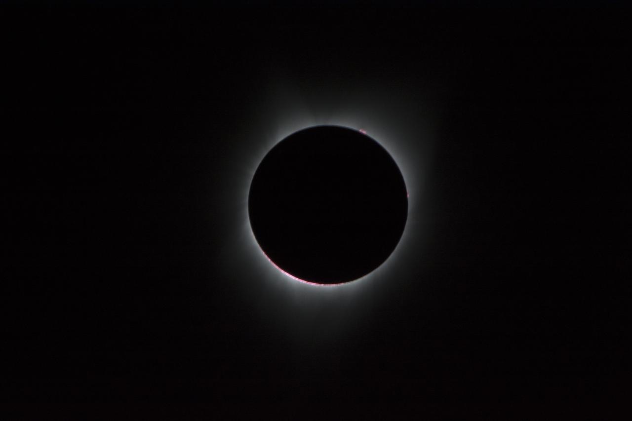Photograph of a total solar eclipse: the Moon covers the Sun and the white corona glows around it, with red prominences at the edge